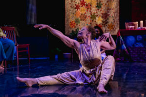 A dancer sits on stage with one leg extended and the other bent, wearing a shimmery jumper and gesturing dramatically to her right.