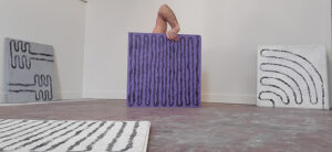An arm bent out the elbow reaches from behind a squiggly set of lines on a purple rectangle. Other squiggly lined-canvases are nearby.
