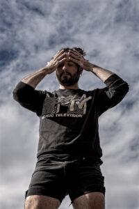 Andrew is standing with hands on their face wearing a shirt and shorts. The photo is taken from below and the backdrop is a cloudy blue sky.