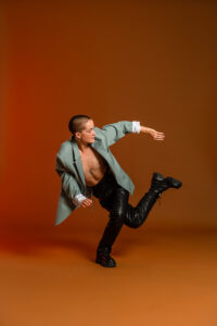 Bobby bends one knee and swings the other leg behind them, twisting to look backward. They are wearing black pants and a gray open jacket. The background is orange.