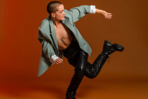 Bobby bends one knee and swings the other leg behind them, twisting to look backward. They are wearing black pants and a gray open jacket. The background is orange.