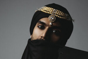 SNJV is close to the camera, dressed in a black head wrap with fabric across the bottom of their face. An ornate piece of jewelry encircles their head and hangs above their face.