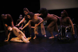 Four standing dancers and two dancers in wheelchairs reach out to a dancer who crouches onstage wearing a mask.