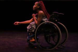 A dancer in a wheelchair is pictured onstage from the side. She leans forward and extends her hand.