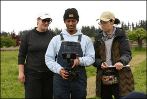 Three people are outside on grass looking at a camera.