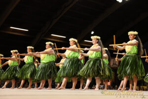 Several dancers perform in unison on a stage bringing two sticks together and extending one foot. They are wearing green grassy skirts and green shirts with flowers in their hair and around the necks.