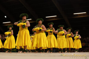 Several dancers in unison and in rows look to the side and bring their arms into their chests. They are wearing yellow and garlands around their heads, necks, and wrists.