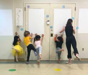 Several children and a teacher stand against a wall and raise their legs behind them.
