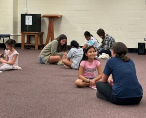 Kids sit across from their adult mentors with their legs criss crossed on a carpet floor.