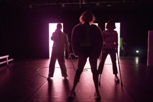 Three dancers stand in a triangle onstage in front of bright pinkish lights.