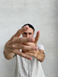 Alessandro stands against a white wall with his hands out in front of him appearing close in the foreground.