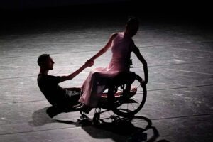 A dancer in a wheelchair holds hands with a dancer sitting on the floor facing her.