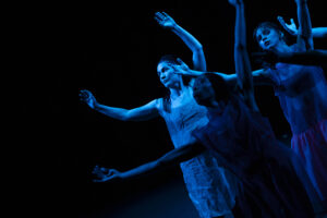Three dancers in blue stage lighting stand and lift their arms.