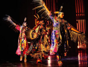 Dancers on a stage wearing regalia with feathers extend their arms so the feathers appear like wings.