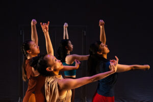 Four dancers onstage face to the side with one arm extended in front of them and one above their heads.