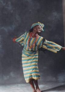 'Funmi dances with her arms outstretched and her head back. She wear green and yellow traditional African dress with red beads and braids in her hair.