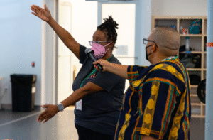 JJ Omelagah, holding a mic for Kayla Hamilton, inside of a Movement Research studio. Kayla is demonstrating a movement for the How We Move cohort with both arms pointing in opposing directions. Both folks are masked.