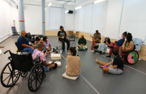 How We Move artists, facilitators, PCAs, and Access Doula gathered in a circle doing access check ins at the studio. Everyone is masked, and there are cordless microphones throughout the space. Folks are surrounded by different chair options, blankets, and cushions, and several people have mobility devices.