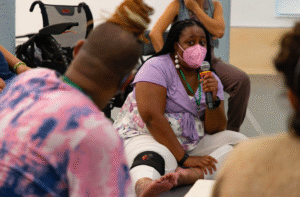 Artist and How We Move facilitator, India Harville, teaching on the floor with a mic. She is surrounded by How We Move artists, PCAs, and Access Doulas.