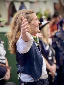 A person with long hair, wearing a white shirt and dark vest, stands outdoors with arms outstretched, surrounded by several people in the background.
