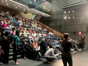 A theater filled with people seated in tiered rows. A few individuals are standing or moving near the stage area. The lighting is bright with several spotlights directed at the stage.
