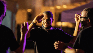 Three individuals are standing and interacting in a warmly lit, indoor or nighttime setting. The background is softly blurred. The central figure’s face is obscured by a blur, while the other two are partially visible in profile. All appear to be engaged in conversation or social interaction.