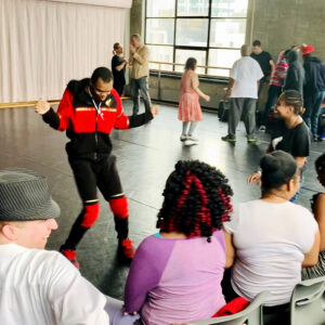 A group of people in a dance studio with a dark floor and concrete walls. Some individuals are dancing or standing, while others are seated on chairs. A large window in the background lets in natural light, and a white curtain hangs on one side of the room.