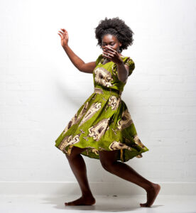 'Funmi wears a green patterned African dress with Afro hear. She steps to the side with her arms gently raised.