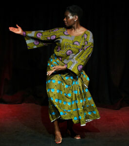 'Funmi wears green patterned traditional African clothing and gestures both hands to the side. A black curtain hangs behind her.