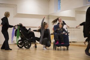 A group of individuals, including those in wheelchairs, are participating in a dance class in a studio with a wooden floor.