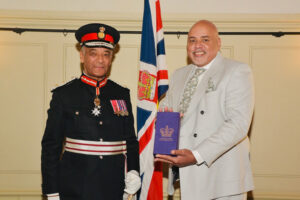 A person in a formal military uniform stands next to another person in a light-colored suit holding an award in front of a flag.