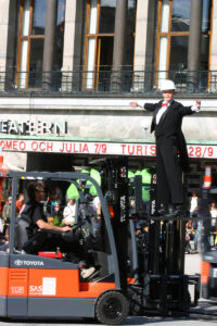 A person in formal attire stands with arms outstretched on the raised forks of a forklift in an outdoor urban setting.