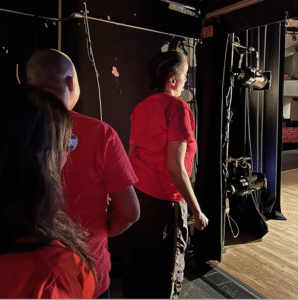 Three individuals wearing red shirts stand backstage. Stage lighting equipment is visible around them, and the audience seating area can be seen in the distance.