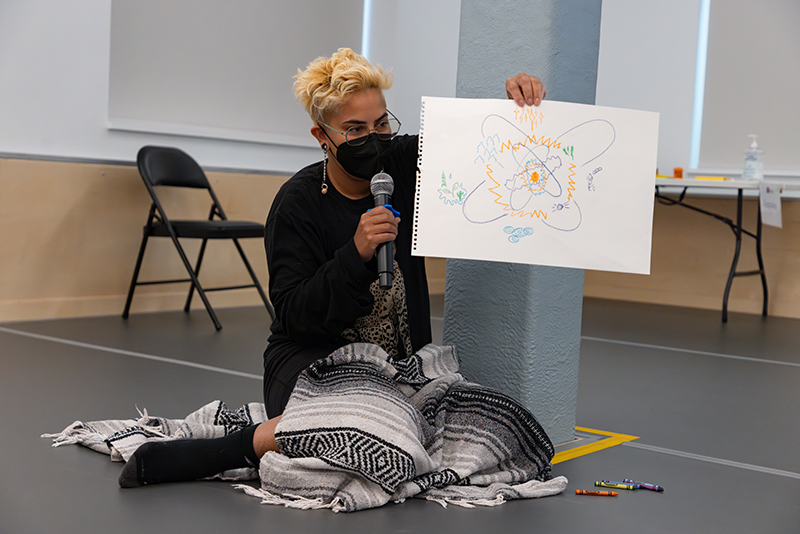 kumari giles, a genderfabulous light brown masked person with short blonde bleached hair with shaved sides and glasses. They are wearing black clothing with a gray/black/white blanket over their lap while sitting on the studio floor, knees bent and presenting an art piece during a workshop while leaning on a gray beam.