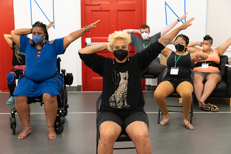 Various masked How We Move participants seated in chairs and mobility devices rehearsing accessible majorette dance in a studio. Everyone’s right arm is bent, with their hand resting on the back of their head, and everyone’s left arm is outstretched. Behind the dancers, there are two red doors and a white wall.