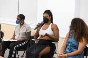 Devin Hill, a masked caramel-skinned Black indigenous person with brown eyes, with black braids wearing a white tank top and black skirt. They’re seated, speaking into a wireless mic during a share back at a Movement Research studio. Beside them are two How We Move PCAs, Tieraney Carter on the left and Barbara Caridad Meulener on the right.