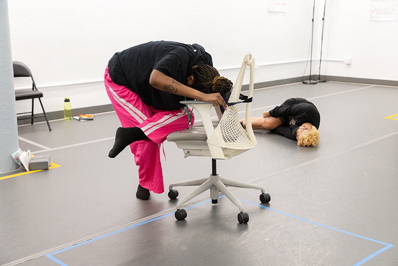 Devin Hill, a masked caramel-skinned Black indigenous person with brown eyes, with black braids wearing a black top and pink pants with white stripes in the studio. They’re standing on their left foot, bent over, leaning on a white chair with their arms resting on the chair arms. Their right leg is bent and resting on the seat of the chair. The floor has been marked with blue tape around them to support their access. In the background is HWM artist, kumari giles, is dancing on the floor.