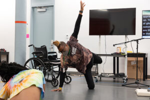 Hector Machado, a masked Afro-Latino brown skin non-binary transfemme with long blonde braids in a bun. She is dancing next to her wheelchair with her right palm and right lower limb touching the studio floor. Her head is down, and her left arm and left lower limb are both outstretched. Beside her is HWM artist, Jackie Robinson.