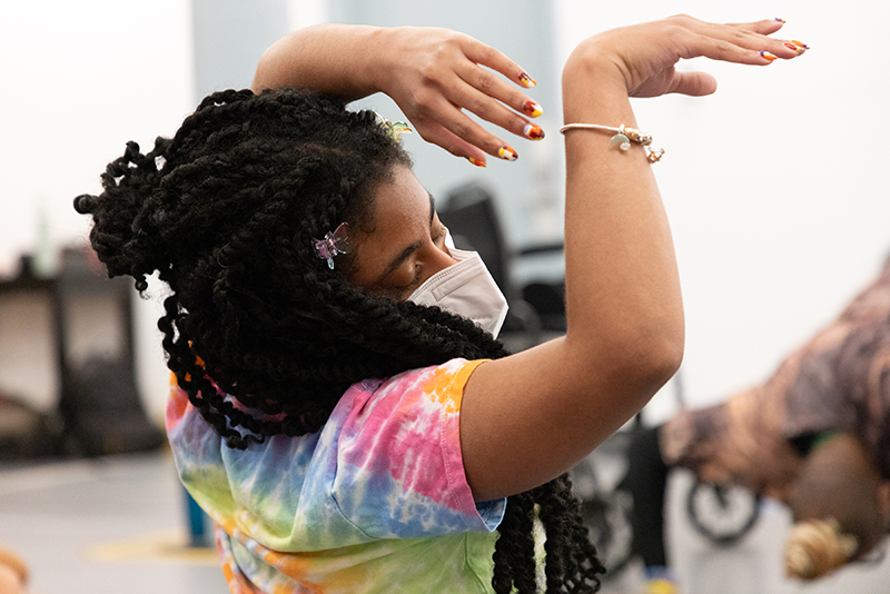 Up close side shot of Jackie Robinson, a masked Black woman with hair styled in twists clipped with butterfly pins wearing a multicolored tie-dye top and fierce nails in How We Move Brand colors. She is dancing in her wheelchair with her head tipped to the side, eyes closed, with hands resting elegantly resting above her head