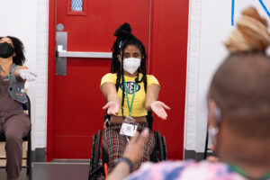 Centered is Jackie Robinson, a masked Black woman with hair styled in twists wearing a yellow crop top and multicolored plaid pants, dancing in her red manual wheelchair. Her arms are extended parallel to her legs with upright palms. She is serving face, looking straight ahead.