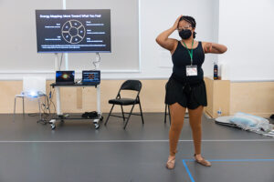Zen Spencer, a masked light-skinned, young Black woman with braids and glasses. She is wearing a black leotard with brown leggings while dancing during her workshop presentation on Energy Mapping. With eyes closed her right hand is cradling her forehead and left arm is raised to shoulder height with hand in front of her shoulder. Three screens showing Zen’s presentation are in the background.