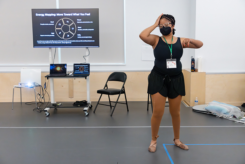 Zen Spencer, a masked light-skinned, young Black woman with braids and glasses. She is wearing a black leotard with brown leggings while dancing during her workshop presentation on Energy Mapping. With eyes closed her right hand is cradling her forehead and left arm is raised to shoulder height with hand in front of her shoulder. Three screens showing Zen’s presentation are in the background.