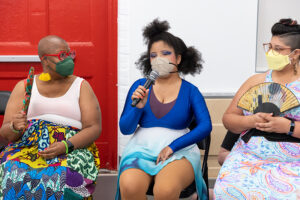 Zen Spencer, a masked, light-skinned, young Black woman with afro puffs, fabulous blue eyeshadow, a blue and purple leotard, and blue, teal, and white wavy dance skirt. She is speaking into a microphone during a share back at Movement Research Studio. Her mom/PCA Aiesha Turman is seated to her left ,and How We Move Staff member Jesenia is seated to her right.