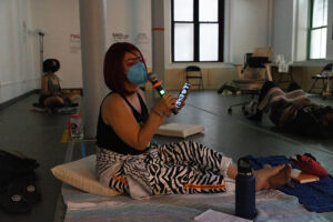Assaleh Bibi is sitting on blankets and cushions with legs stretched out and crossed at the ankle. Xe is wearing a hot pink wig and has hot pink feather eyelashes. Xe is holding a wireless mic and phone as xe facilitates a guided meditation in a dimly lit dance studio. Two other participants are lying down and seated in the background.