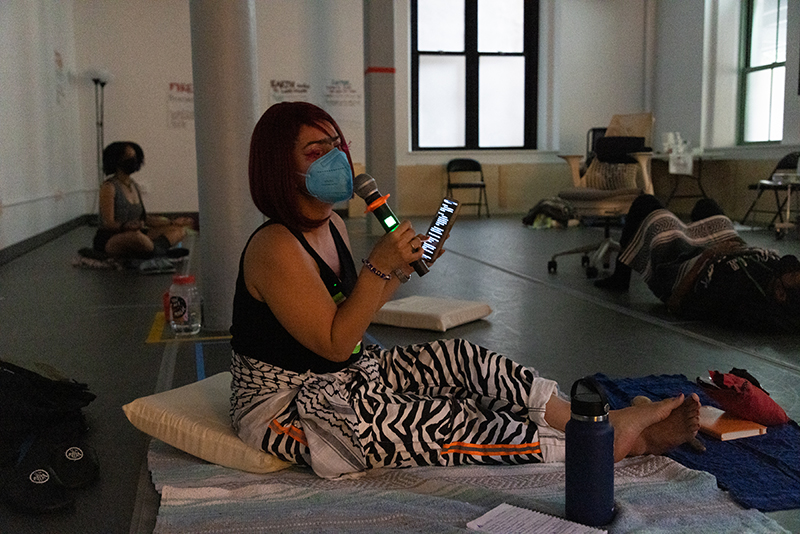 Assaleh Bibi is sitting on blankets and cushions with legs stretched out and crossed at the ankle. Xe is wearing a hot pink wig and has hot pink feather eyelashes. Xe is holding a wireless mic and phone as xe facilitates a guided meditation in a dimly lit dance studio. Two other participants are lying down and seated in the background.