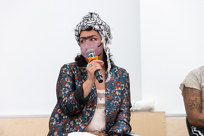 Assaleh Bibi speaks into a microphone while sitting in a chair. Xe is wearing a blue jacket with a multicolored floral design, a pale pink shirt, a pink mask, sparkly pink eyeshadow, and a black and white keffiyeh. There is a white wall behind xem.