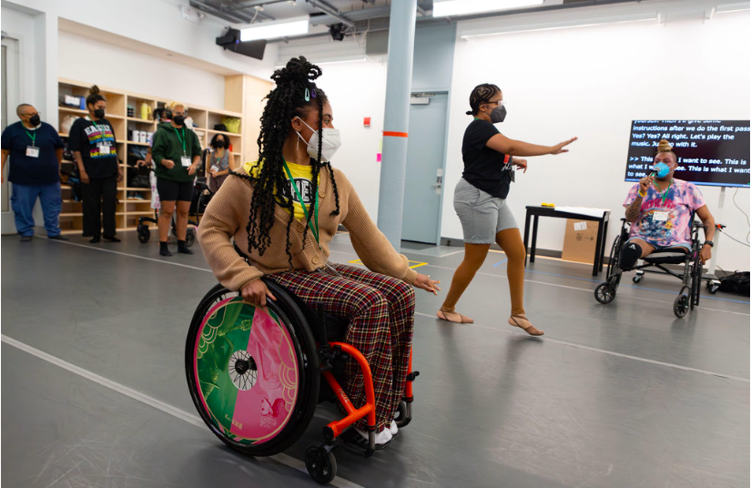 Jackie Robinson is dancing in her wheelchair with left hand pointing outward, and Zen Spencer is strutting with right arm lifted in front of her and left arm out to her side for balance. Other How We Move attendees (JJ Omelagah, Dev Hill, kumari giles, India Harville, and Jazz Goldman) observe on the sidelines. Hector Machado encourages the dancers from the front of the room with the CART screen behind her.
