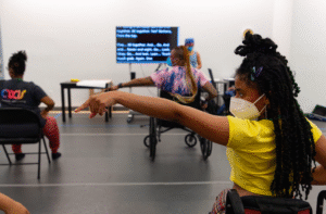 How We Move artists rehearsing majorette dance in the studio. Featured are Hector Machado, Jackie Robinson, and Kayla Hamilton. Everyone is masked with their left arm and index finger pointing outward to the side, and their right hand on their hip. The shot is taken from behind the dancers. In the front, captions are being shared on a large TV screen.