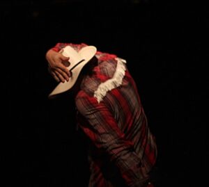 The back of a dancer in a cowboy shirt and hat, one hand looped over the hat. The background is black.