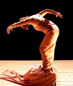 A dancer onstage stands on a box wearing a long pink dress that wraps around her legs and the box. She bends over with arms extended out.